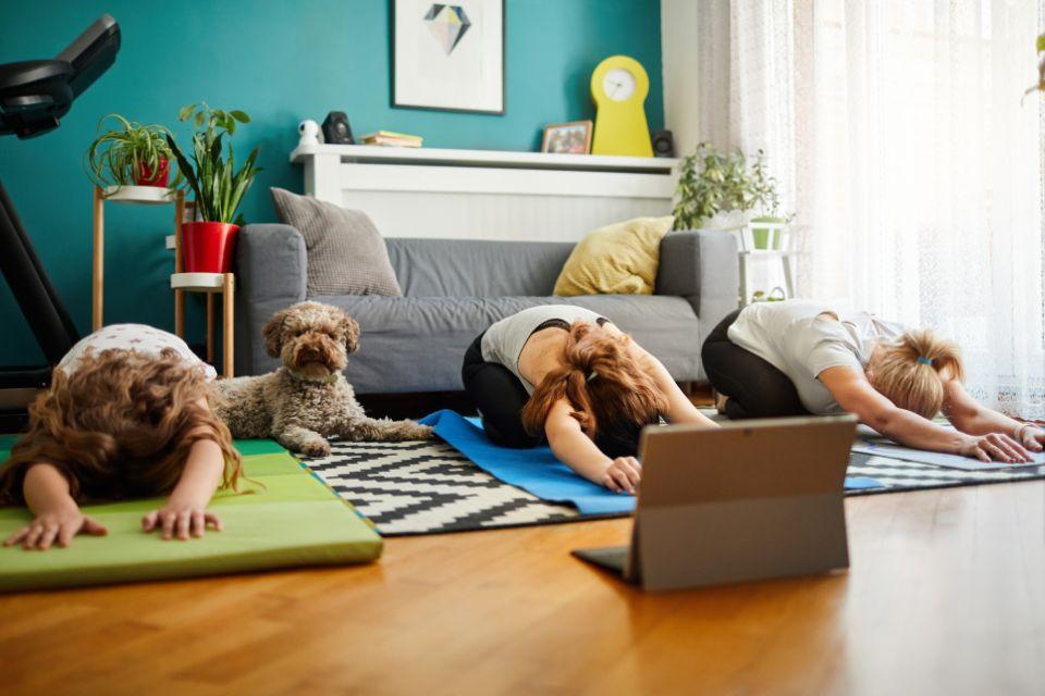 A mother and her two daughters doing child's pose in front of a laptop in their living room while the family dog watches frmo behind