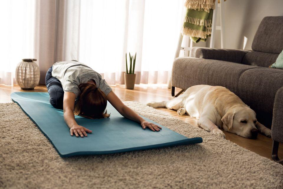 A woman in child's pose on a yoga mat next to her sleeping dog, who is pressed against the couch