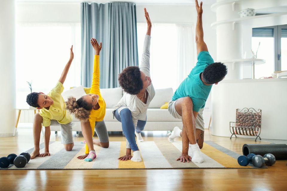 A mother, father, and their two kids doing a runner's twist lunge on their living room carpet with weights and a rolled up yoga mat on the floor in front of them