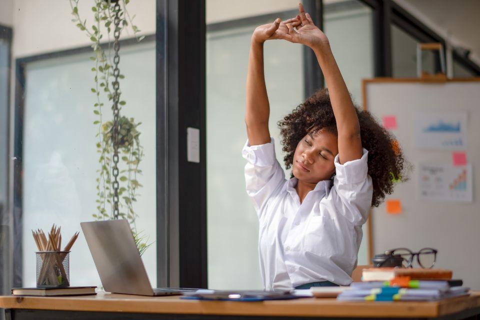 A professional woman stretching her arms and back at her work desk in front of a laptop