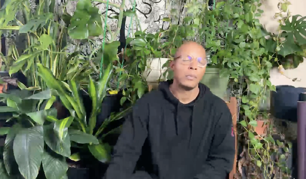 Ompractice teacher Dominic Stanley sitting with eyes closed in front of plant wall
